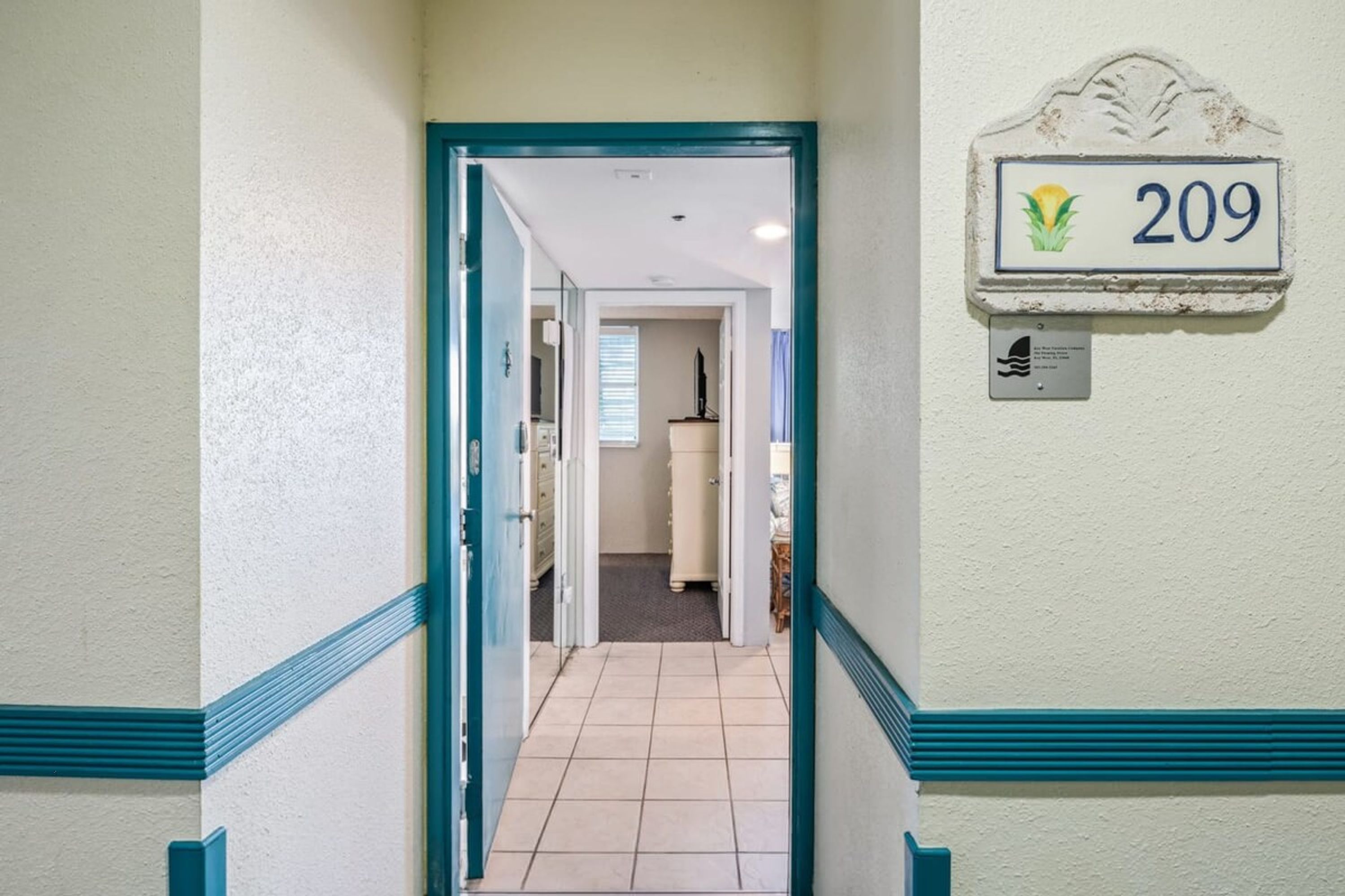 Um corredor com uma porta que dá para um quarto, uma placa com o número 209 na parede, e uma pequena placa logo abaixo.
