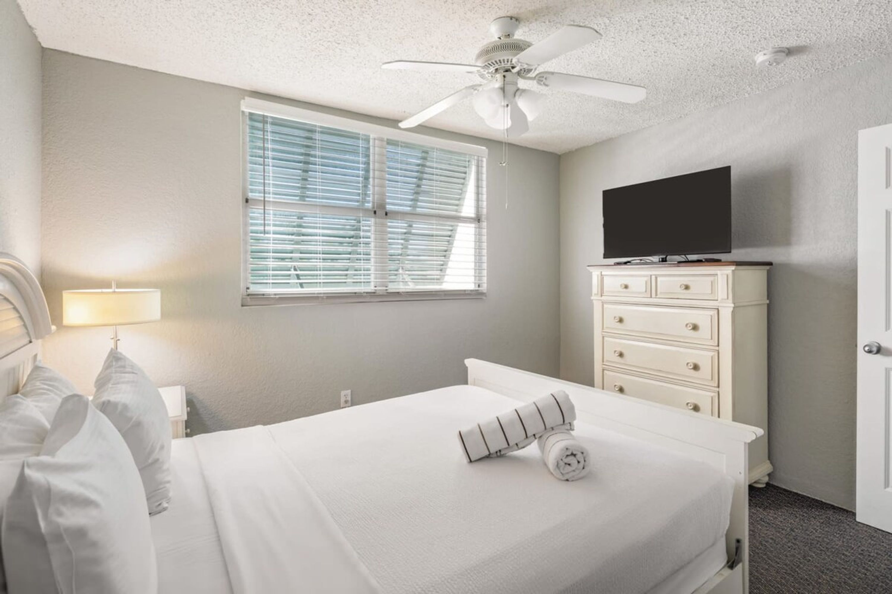 Habitación de hotel con cama, televisión en una cómoda, ventilador de techo y ventana con persianas.