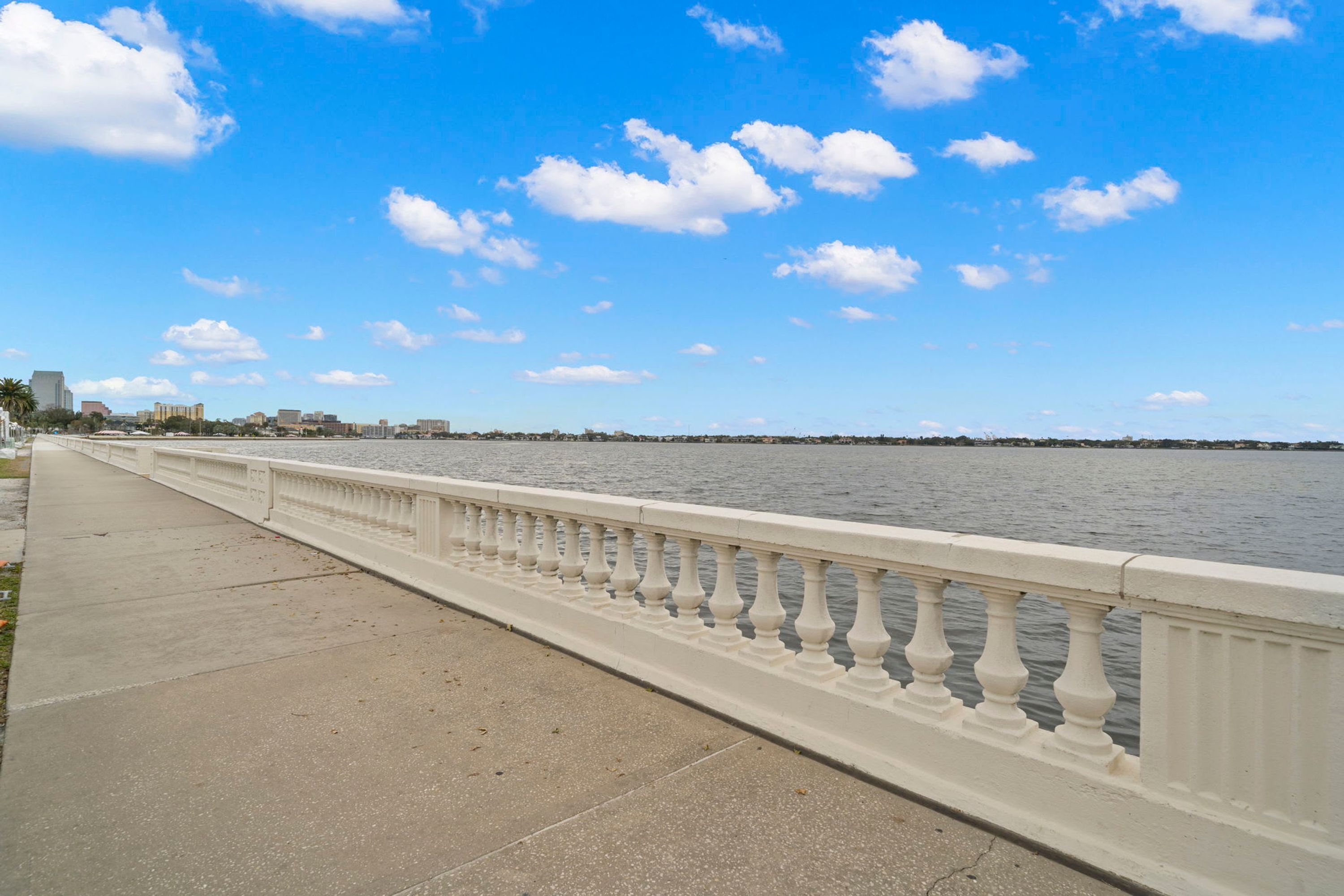A waterfront promenade with a white balustrade, a concrete walkway, and a calm body of water under a clear sky.