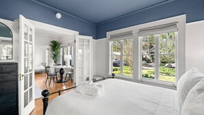 Una camera da letto con un letto grande, biancheria da letto bianca, una scrivania con una sedia e vista sull'esterno attraverso ampie finestre.
