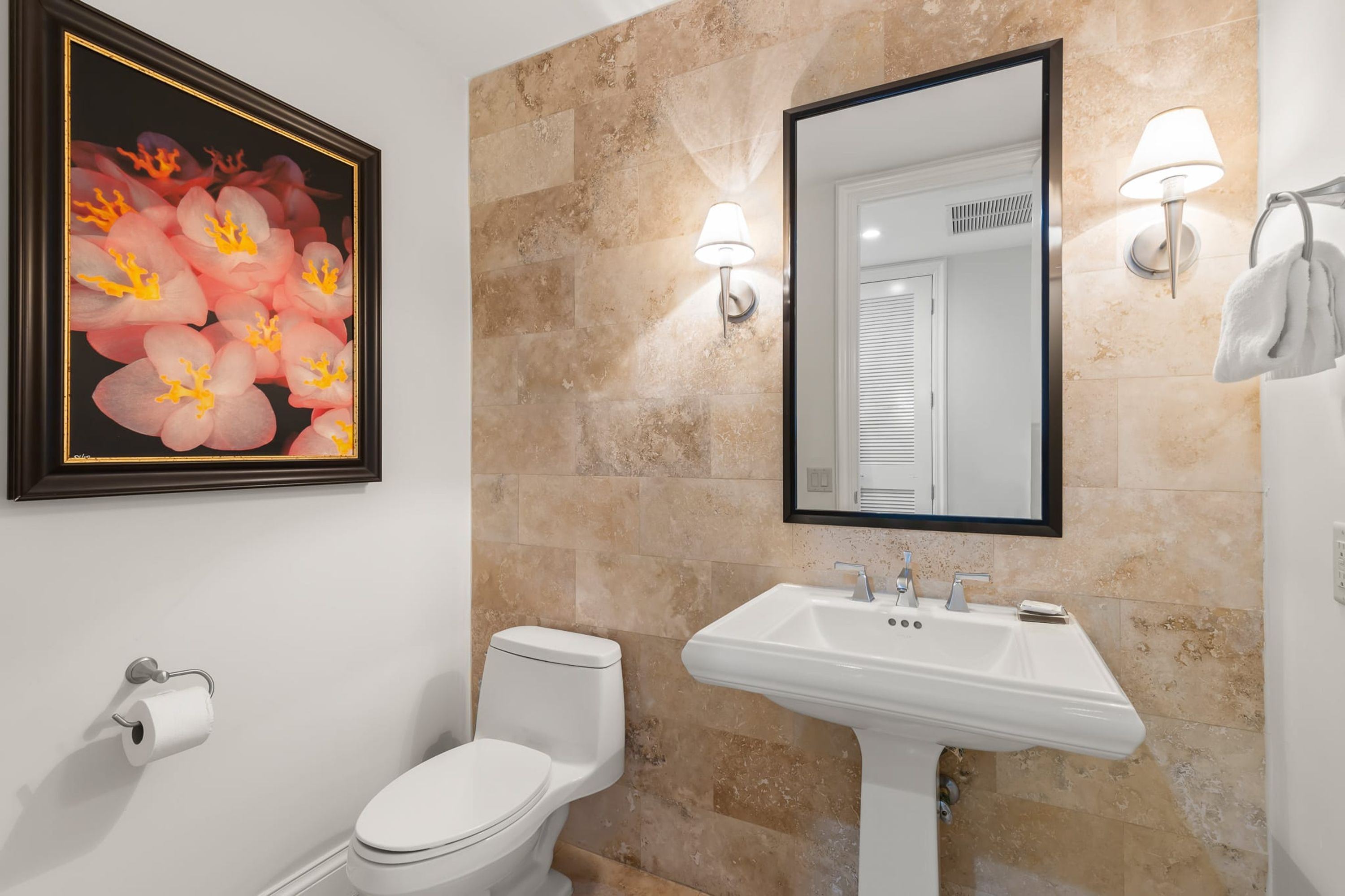 Baño con inodoro, lavabo, una obra de arte floral enmarco y un espejo con luces empotradas.
