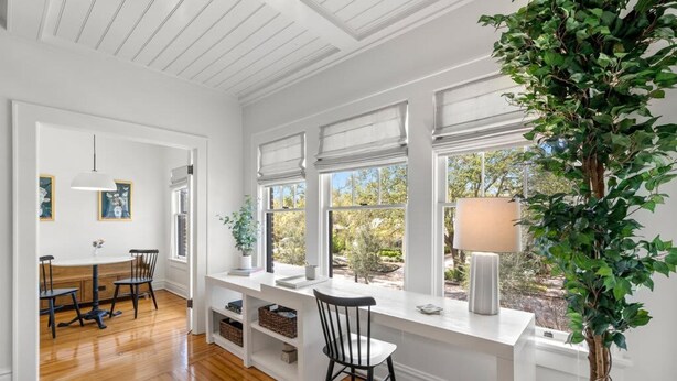 Una habitación luminosa con pisos de madera, paredes blancas y ventanas amplias. Cuenta con una mesa de comedor con sillas, un banco blanco y una planta en maceta.