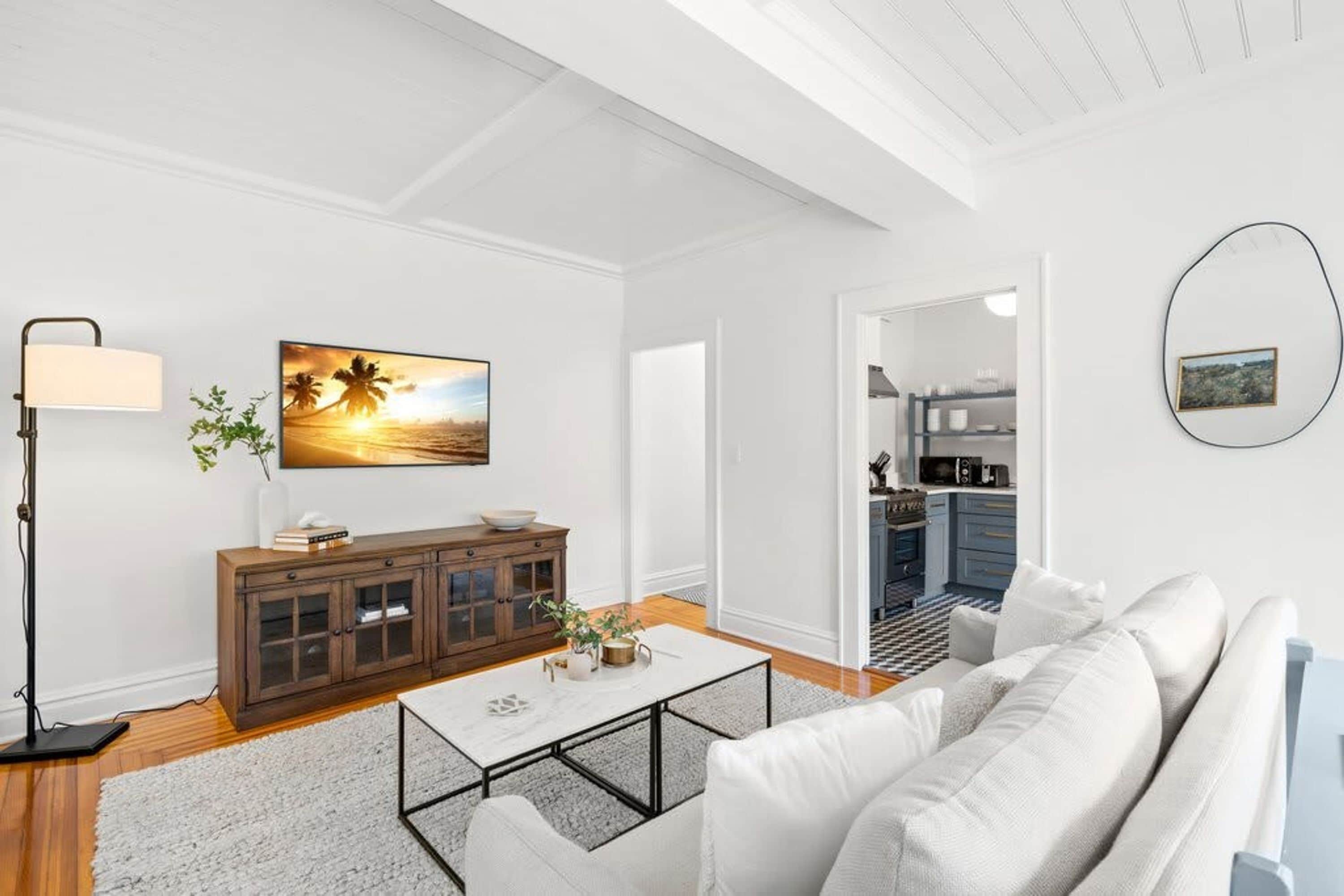 A modern living room with a white sofa, a coffee table, and a painting of a sunset. There is a wooden cabinet and a floor lamp. A kitchen is visible through an open doorway.