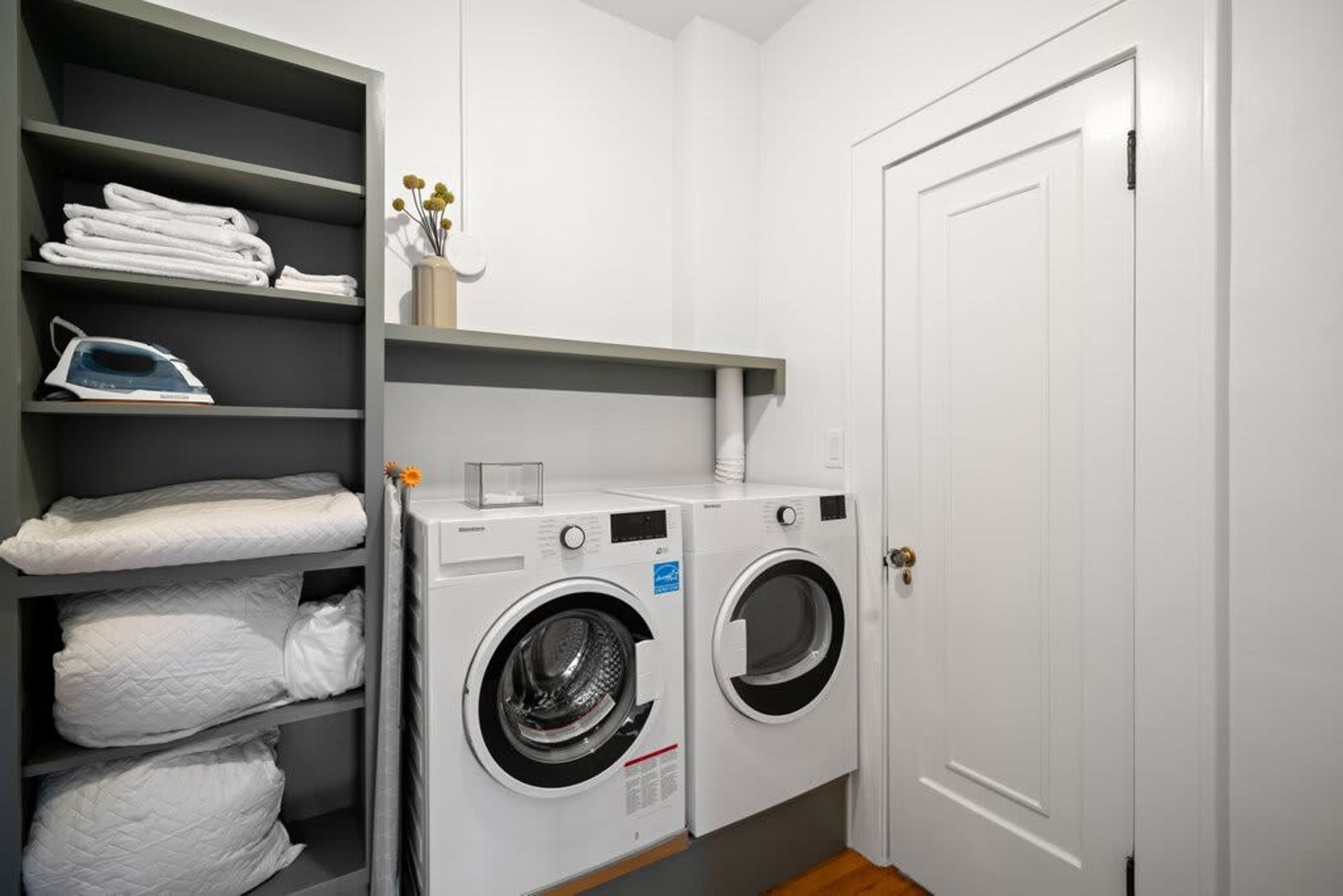 A laundry room with two front-loading washing machines, a shelf with folded towels, and an ironing board.