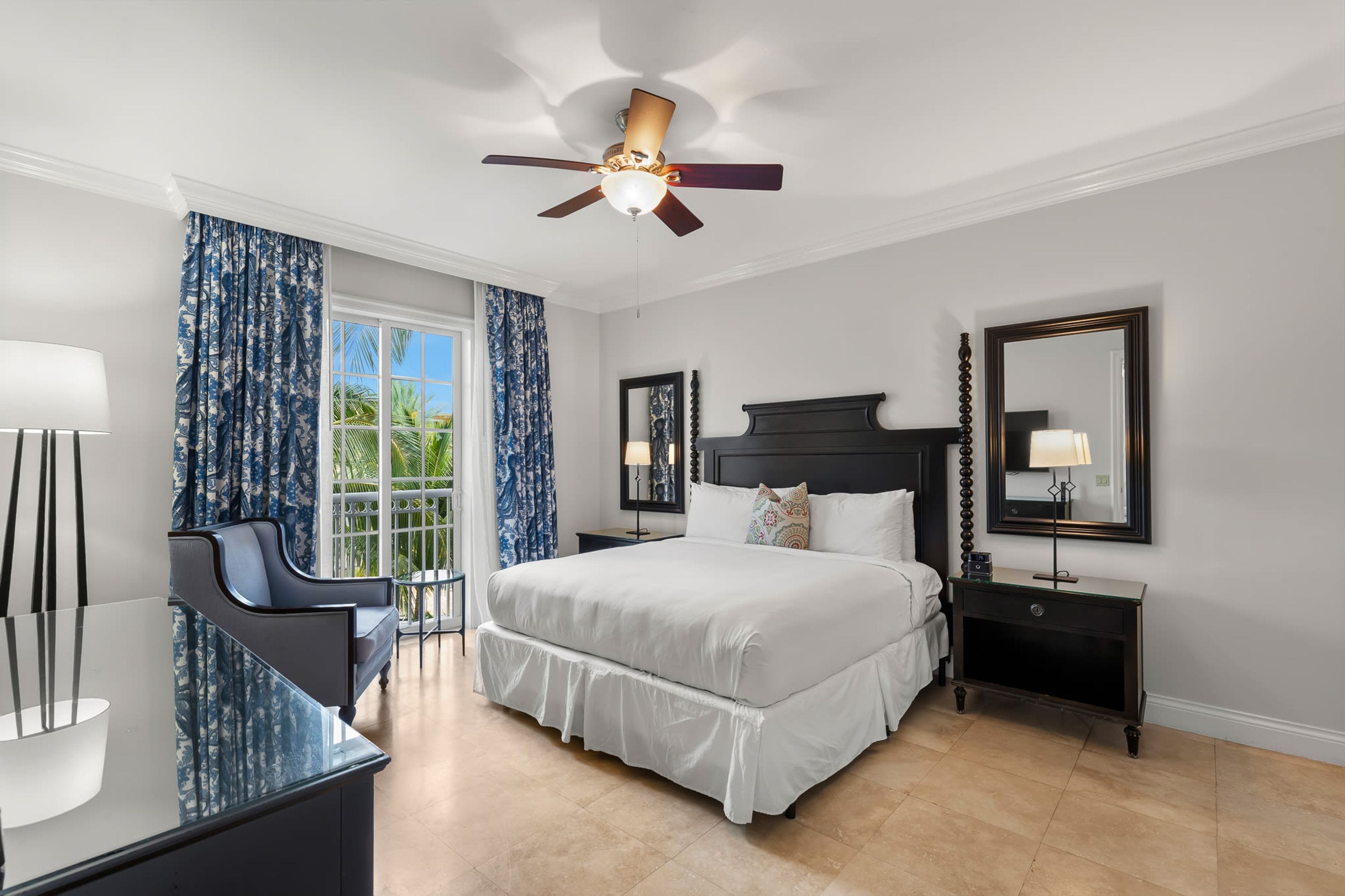 A bedroom with a bed, a desk, a chair, a ceiling fan, and a large window.