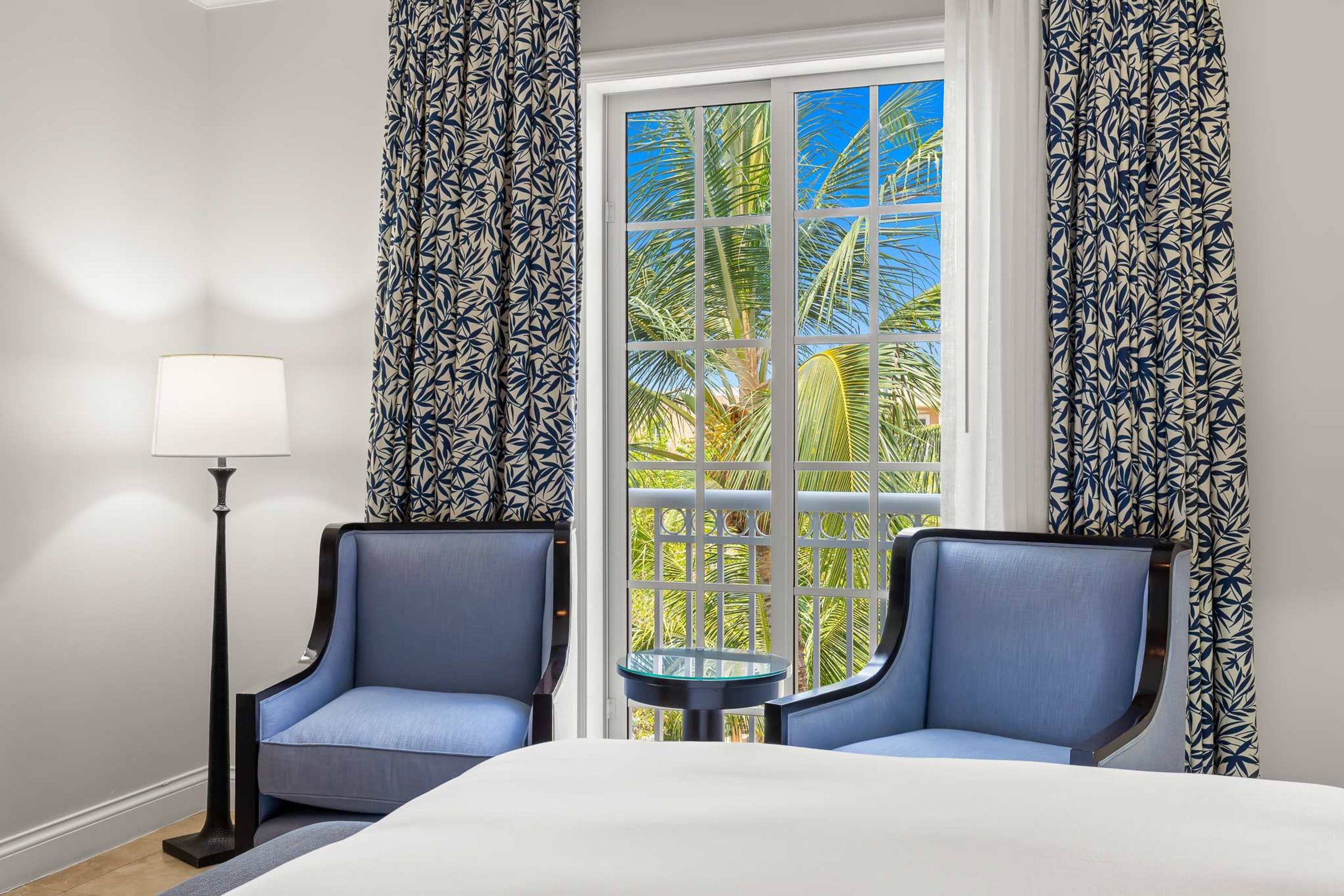 A hotel room with a bed, two armchairs, a small table, a floor lamp, and a window with a view of palm trees.