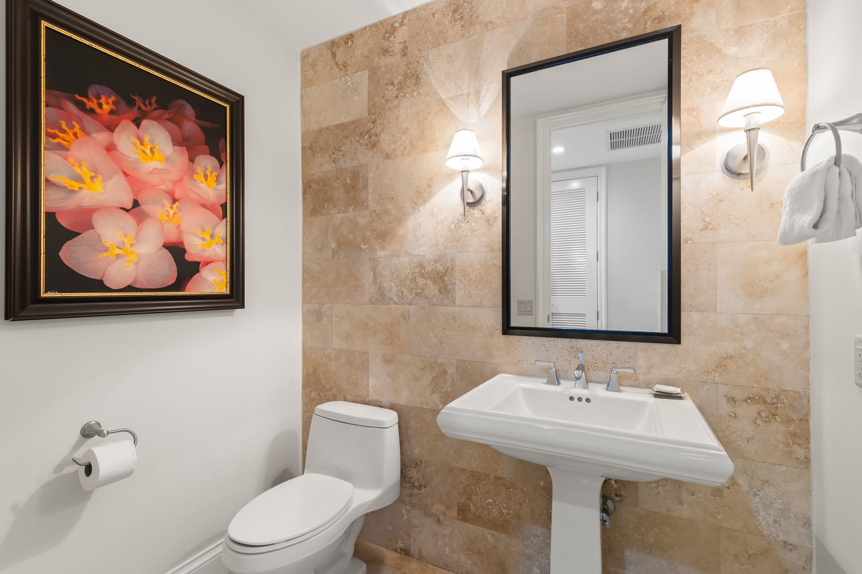 A bathroom with a toilet, sink, framed floral artwork, and wall-mounted lights.