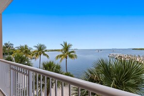 A balcony with a view of a calm sea, palm trees, and a dock with boats.
