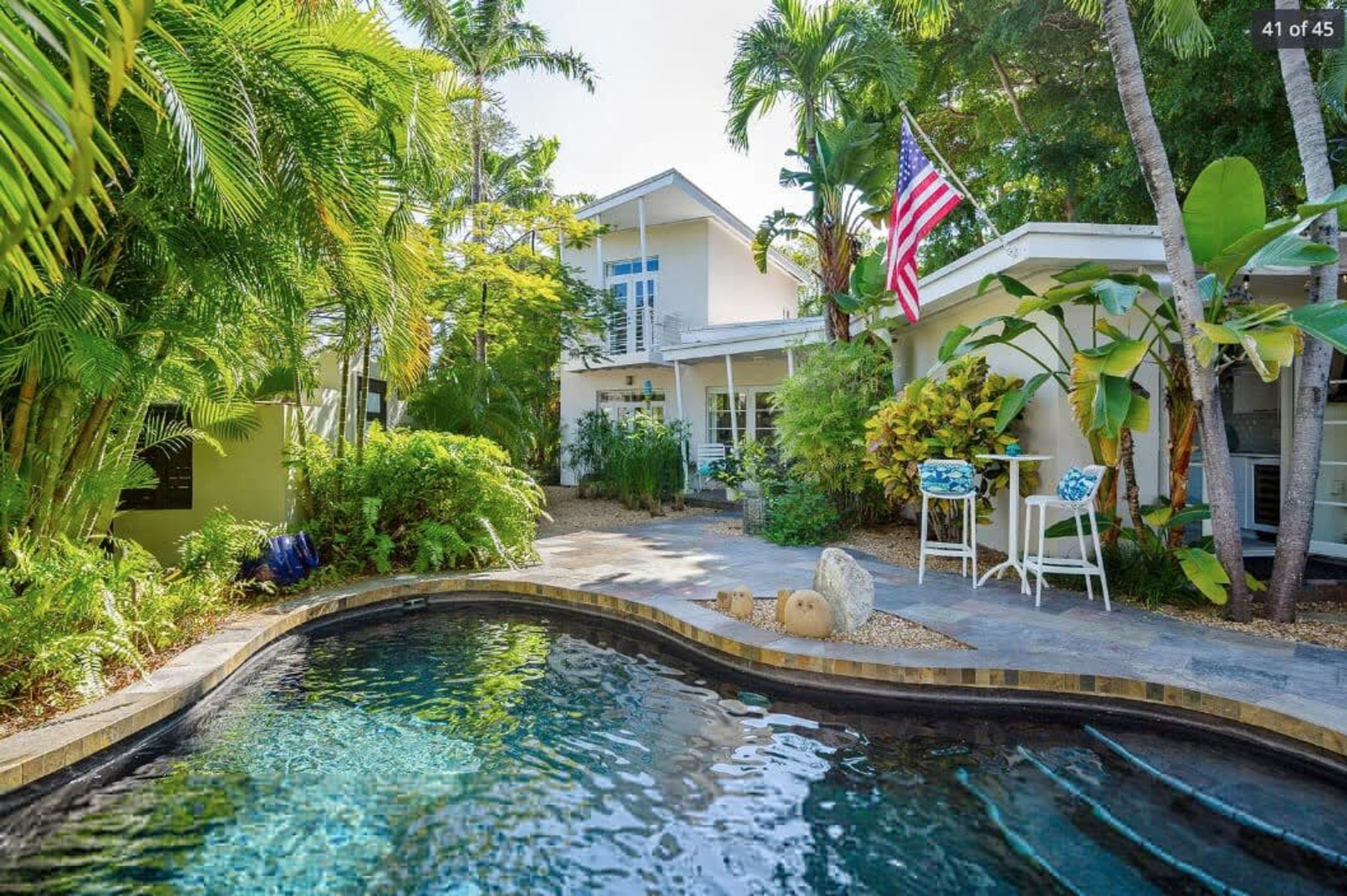 A pool area with a house in the background, surrounded by lush greenery and an American flag.