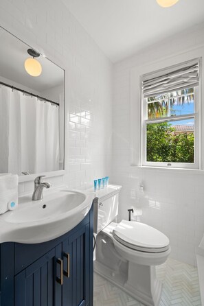 A bathroom with a white sink, dark blue vanity, white toilet, and a window with a view of greenery. - NEW The Adaline 4 Updated Hyde Park Apt (Tampa)