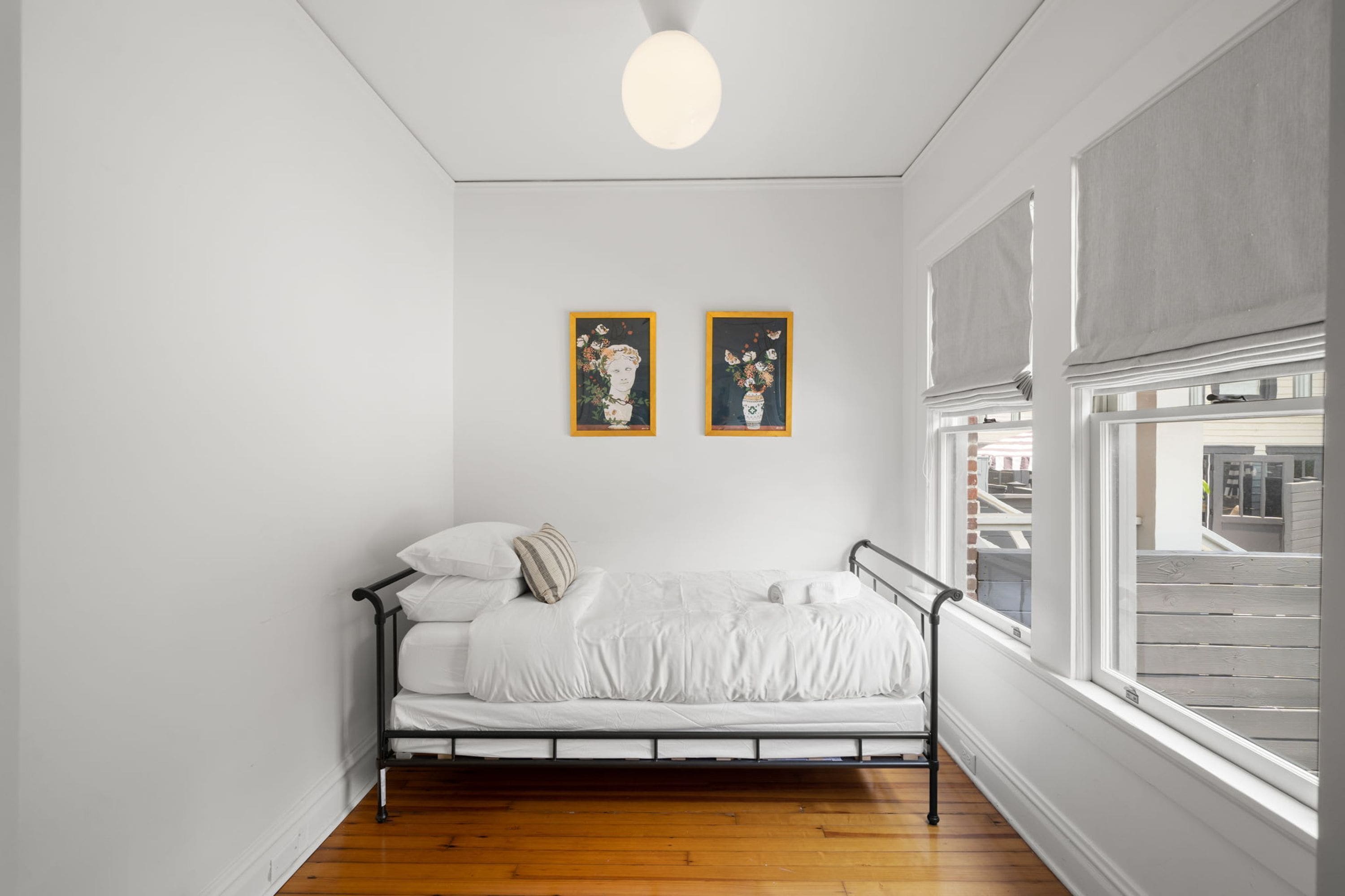 A single bed in a room with white walls and wooden flooring. There are two framed artworks on the wall, a window with blinds, and a ceiling light.