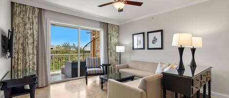 A spacious living room with a sofa, a coffee table, a TV mounted on the wall, a ceiling fan, and a view of the outdoors through a sliding glass door.