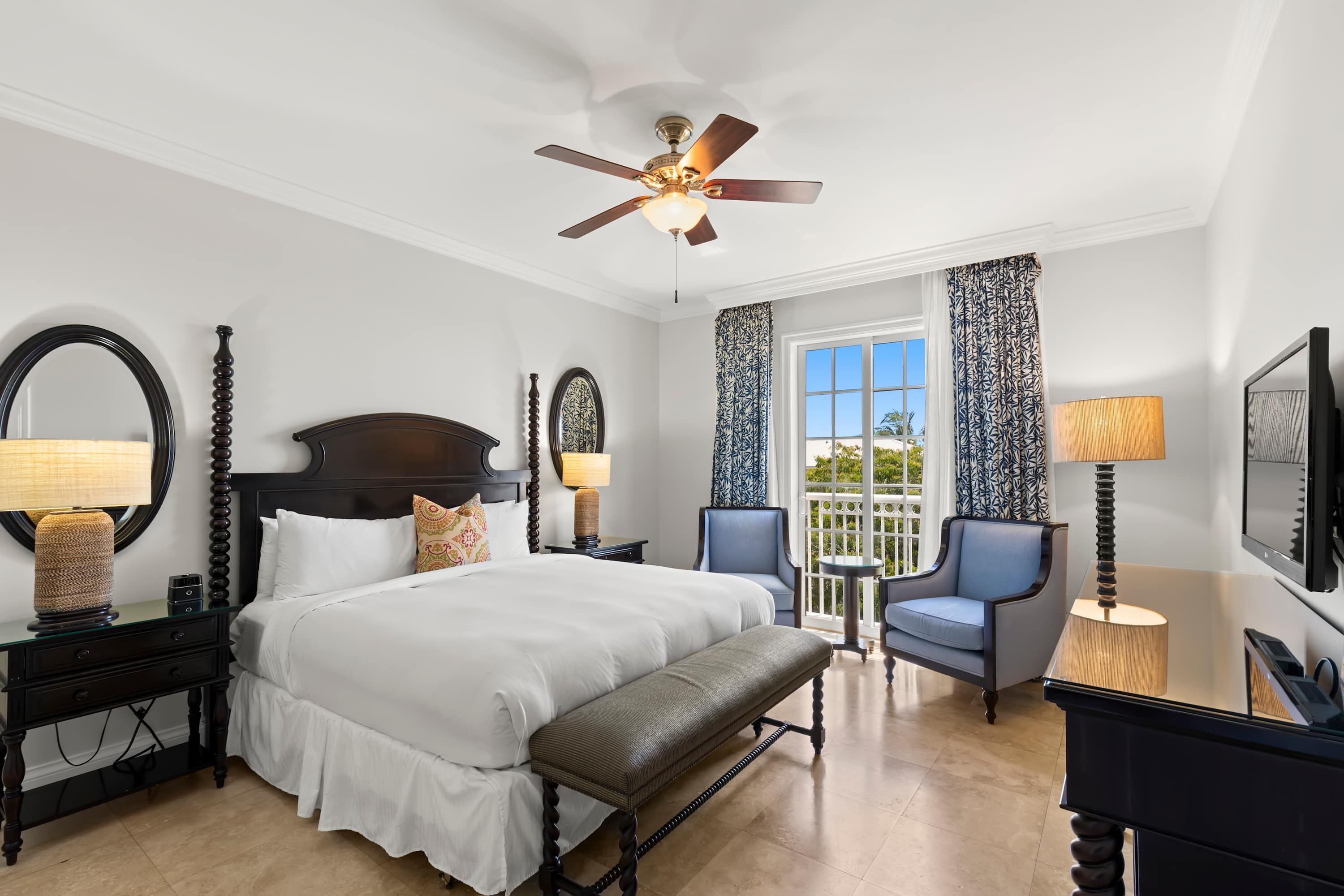A hotel room with a large bed, a bench, a desk, two chairs, a ceiling fan, and a large window with curtains.