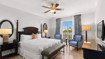 A hotel room with a large bed, a bench, a desk, two chairs, a ceiling fan, and a large window with curtains.