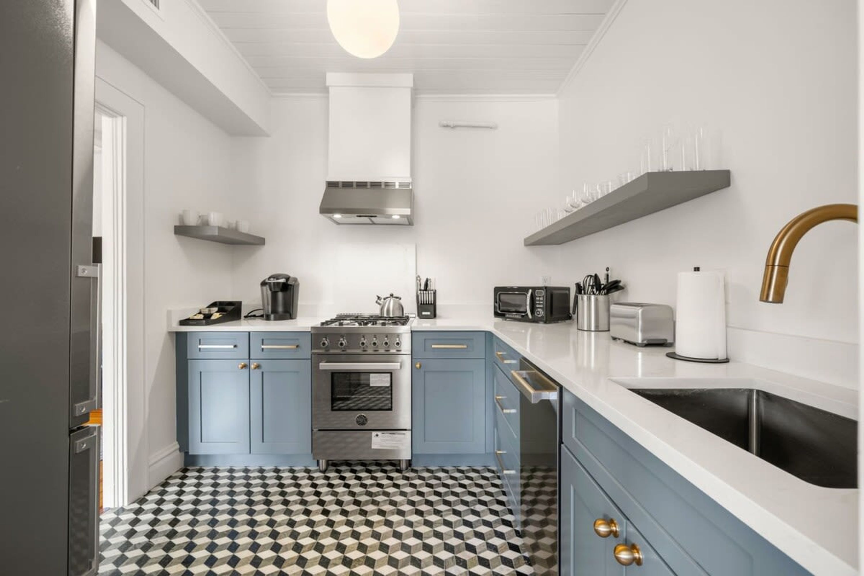 A compact kitchen with a central stove, microwave, and sink. There are cabinets with gold handles and a patterned floor.