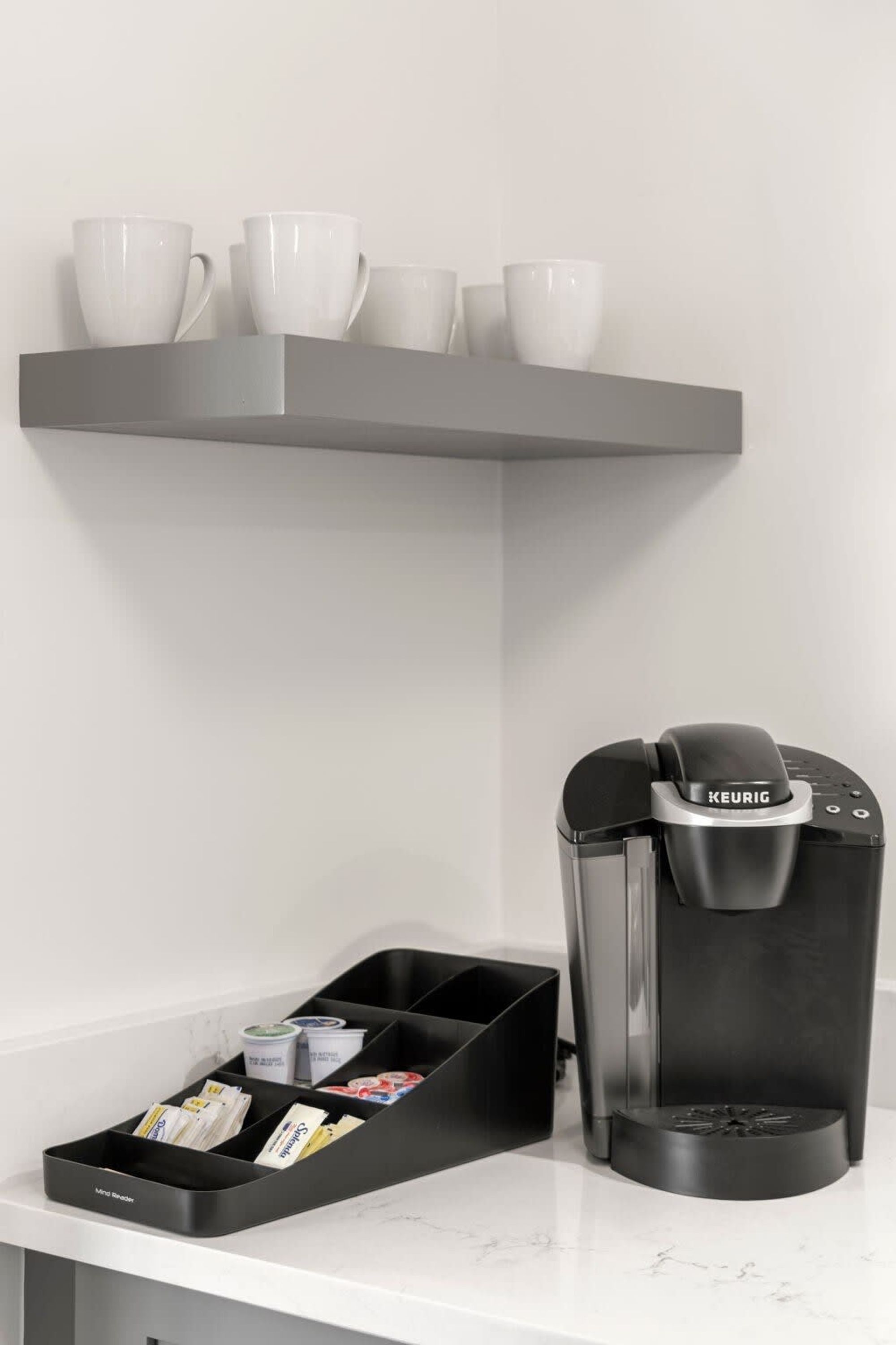 Una cafetera marca Keurig, un dispensador de cápsulas de café y un juego de tazas blancas sobre una estantería.