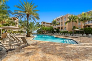 Relax by the clear pool surrounded by lounge chairs and umbrellas, with palm trees and multi-story buildings in the background.