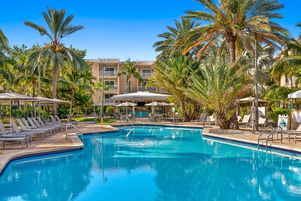 Relax by the resort's clear pool surrounded by lounge chairs and umbrellas, with stunning views of the surrounding buildings and lush palm trees.