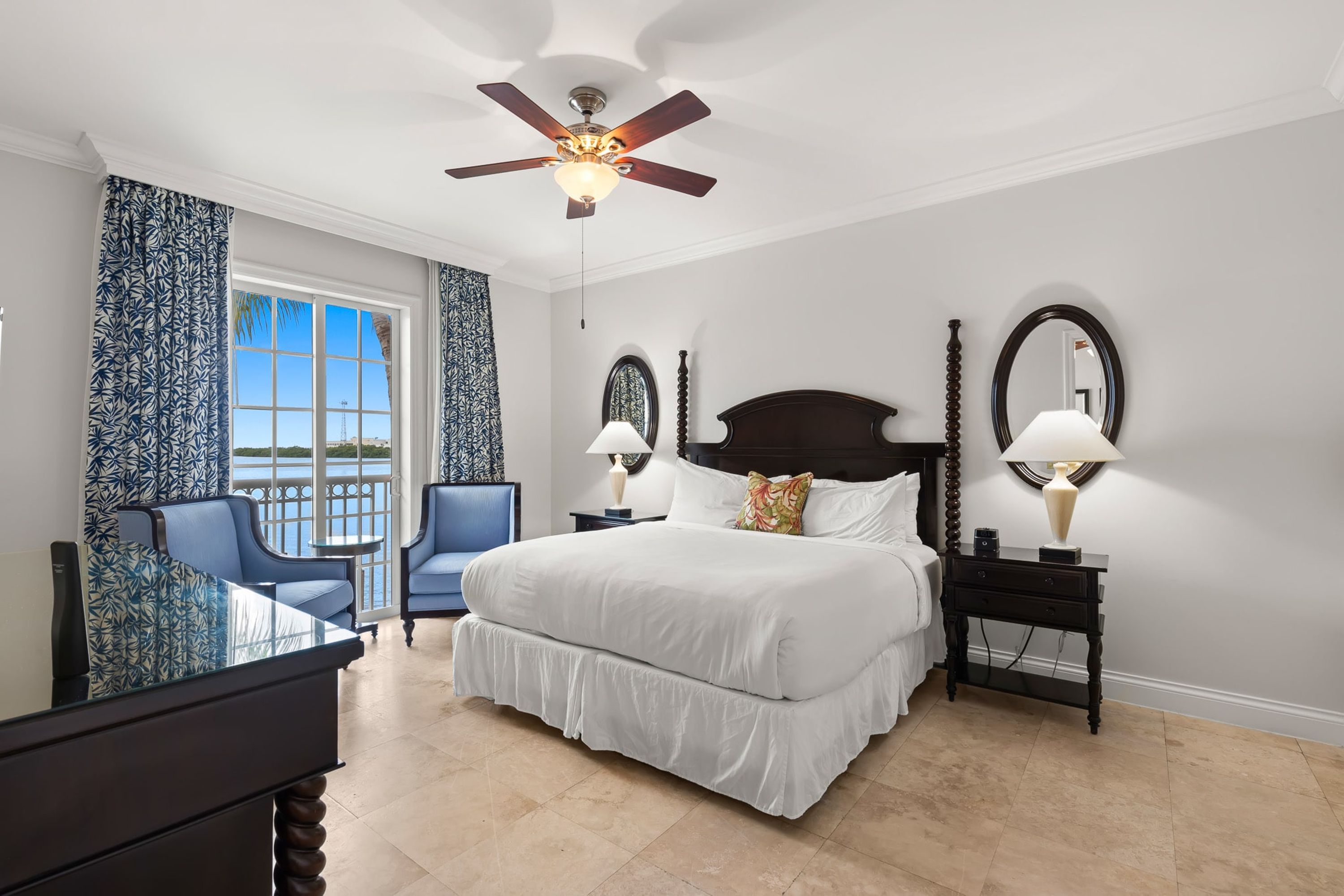 A bedroom with a large bed, two bedside tables with lamps, a ceiling fan, a desk with a chair, and a view of the outdoors through a window.