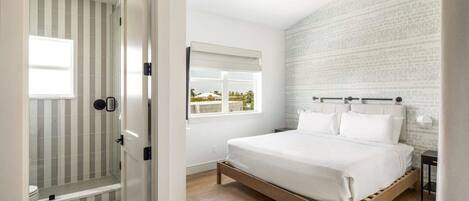A bedroom with a wooden bed, a window with blinds, a ceiling fan, and a shower area with striped tiles.
