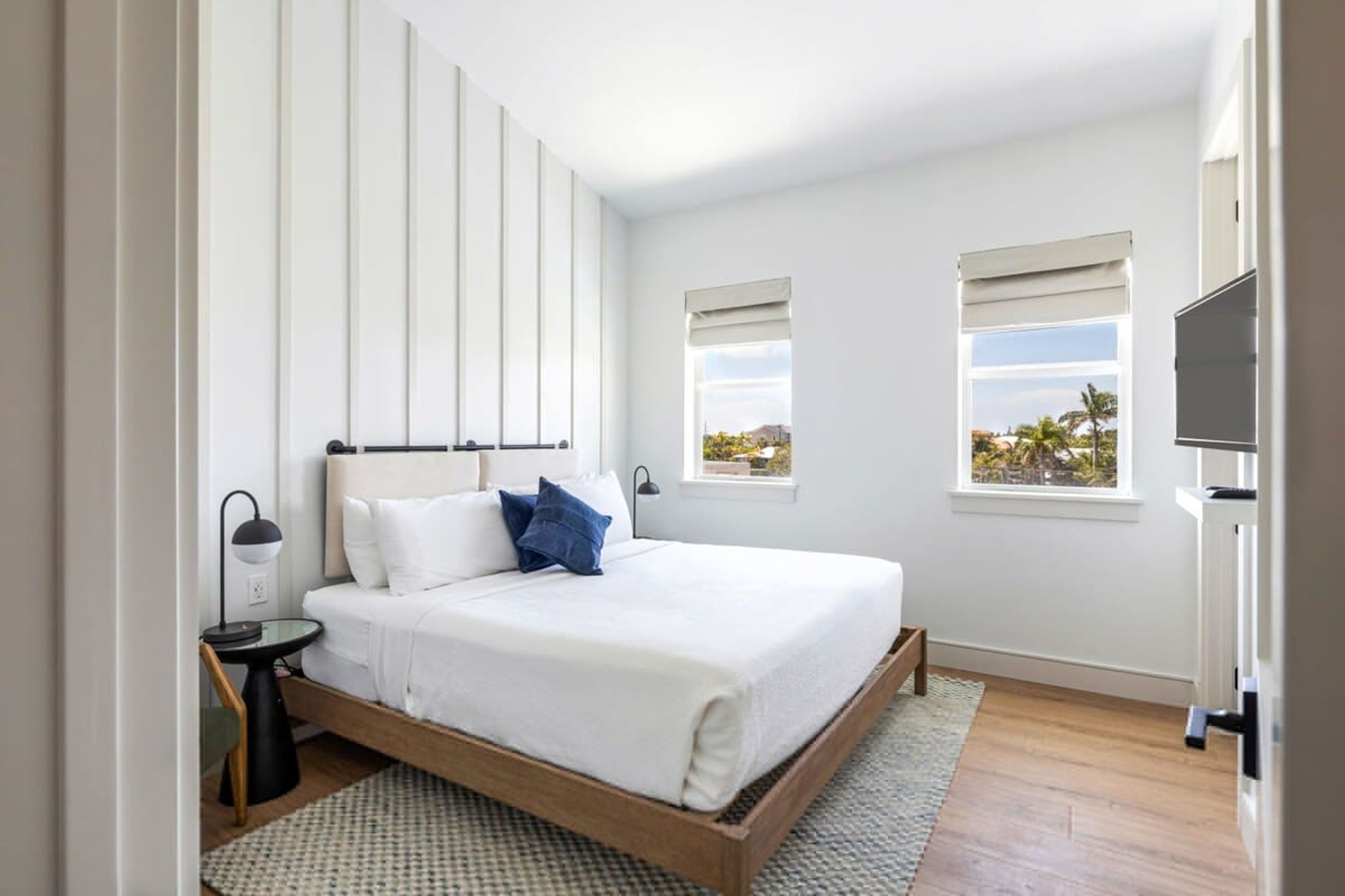 A modern bedroom with a wooden bed, white walls, and two windows with blinds.