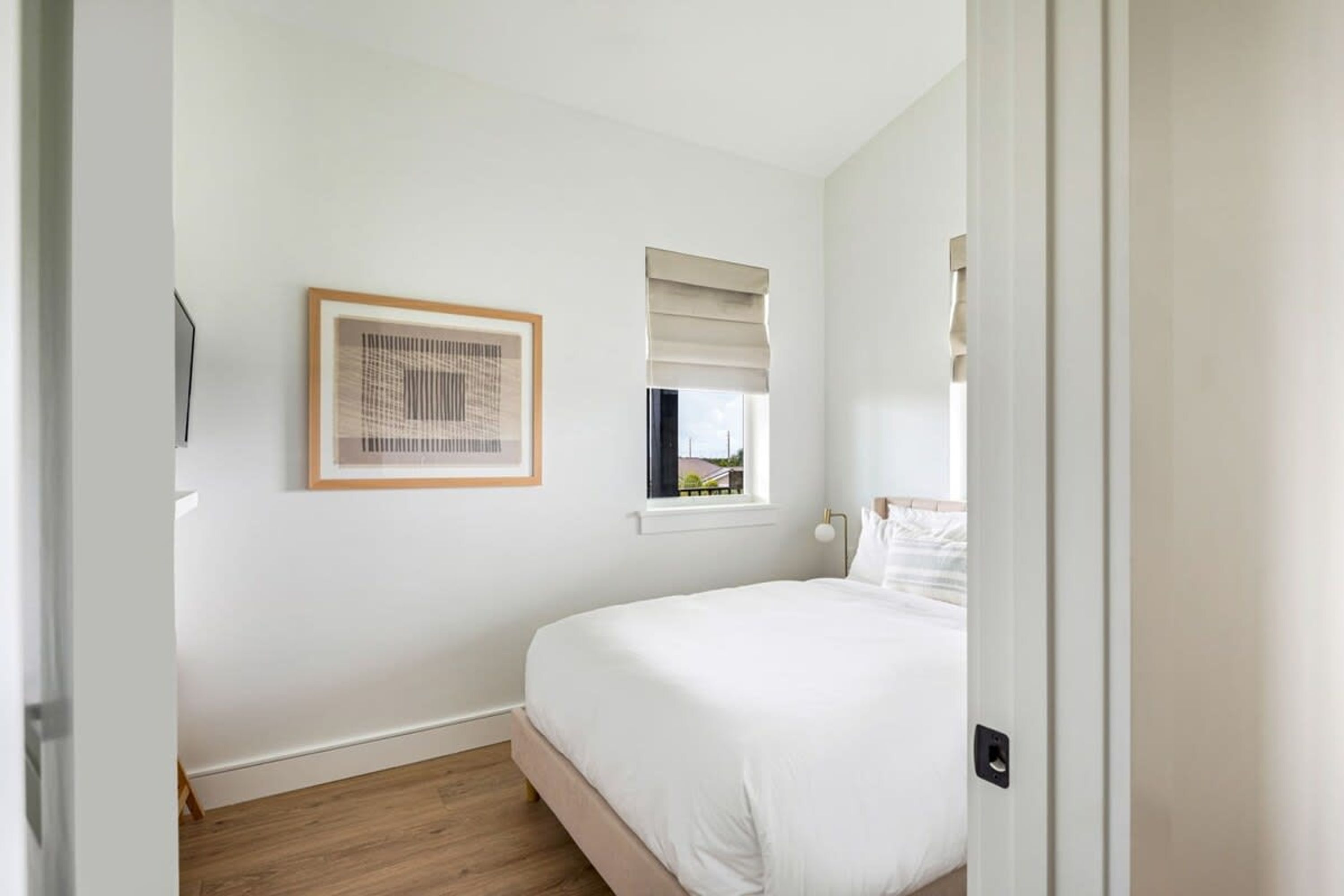 Une chambre à coucher avec un lit, une fenêtre avec des stores, un tableau encadré et une porte.