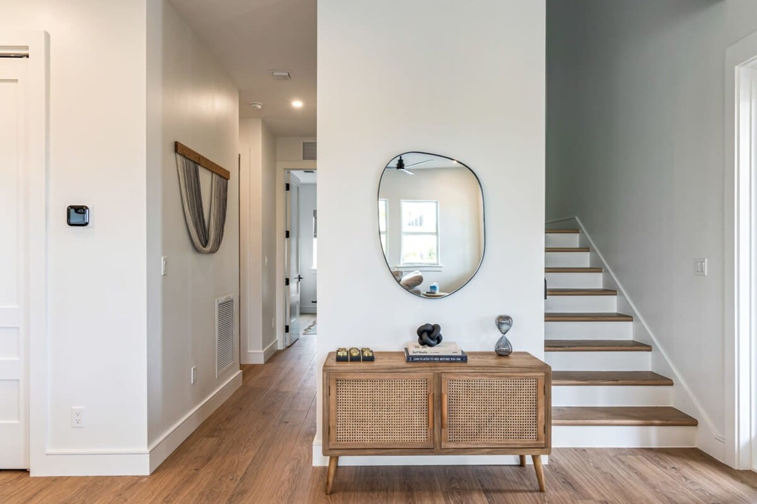 Un couloir avec un plancher en bois, un mur blanc, un miroir rond et une console en rotin ornée de livres et d’objets décoratifs.