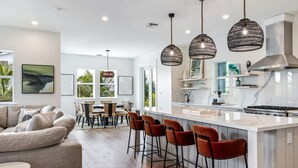 A modern kitchen with a central island, bar stools, pendant lights, and a dining area with a sofa and chairs.