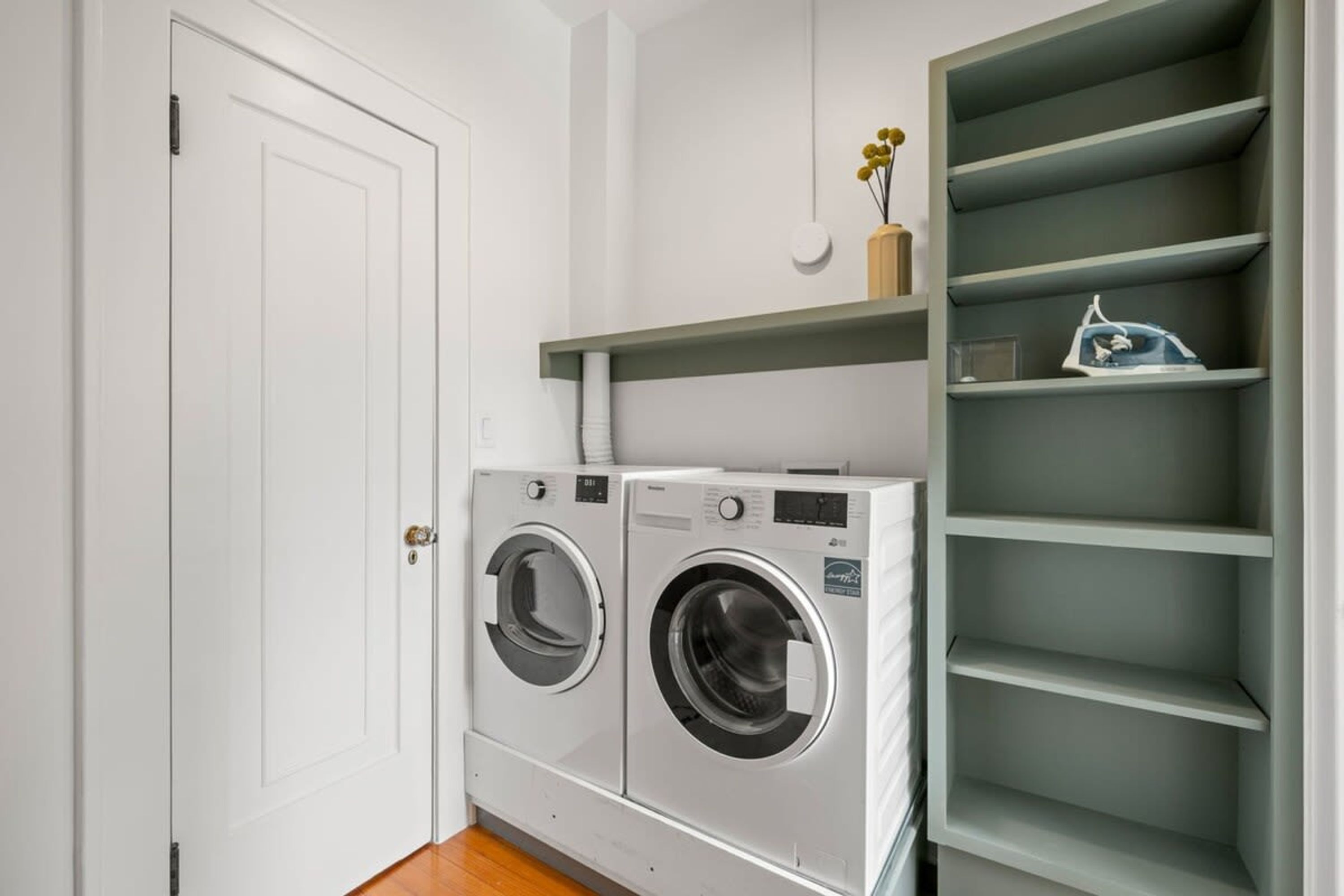 A laundry room with two front-loading washing machines and a green shelving unit above them.