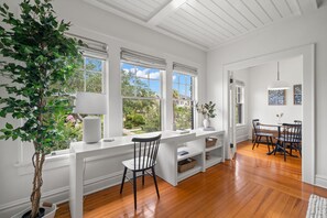 A bright room with a white desk, a chair, and a potted plant. There are windows with blinds, a wooden floor, and a view of the outside.