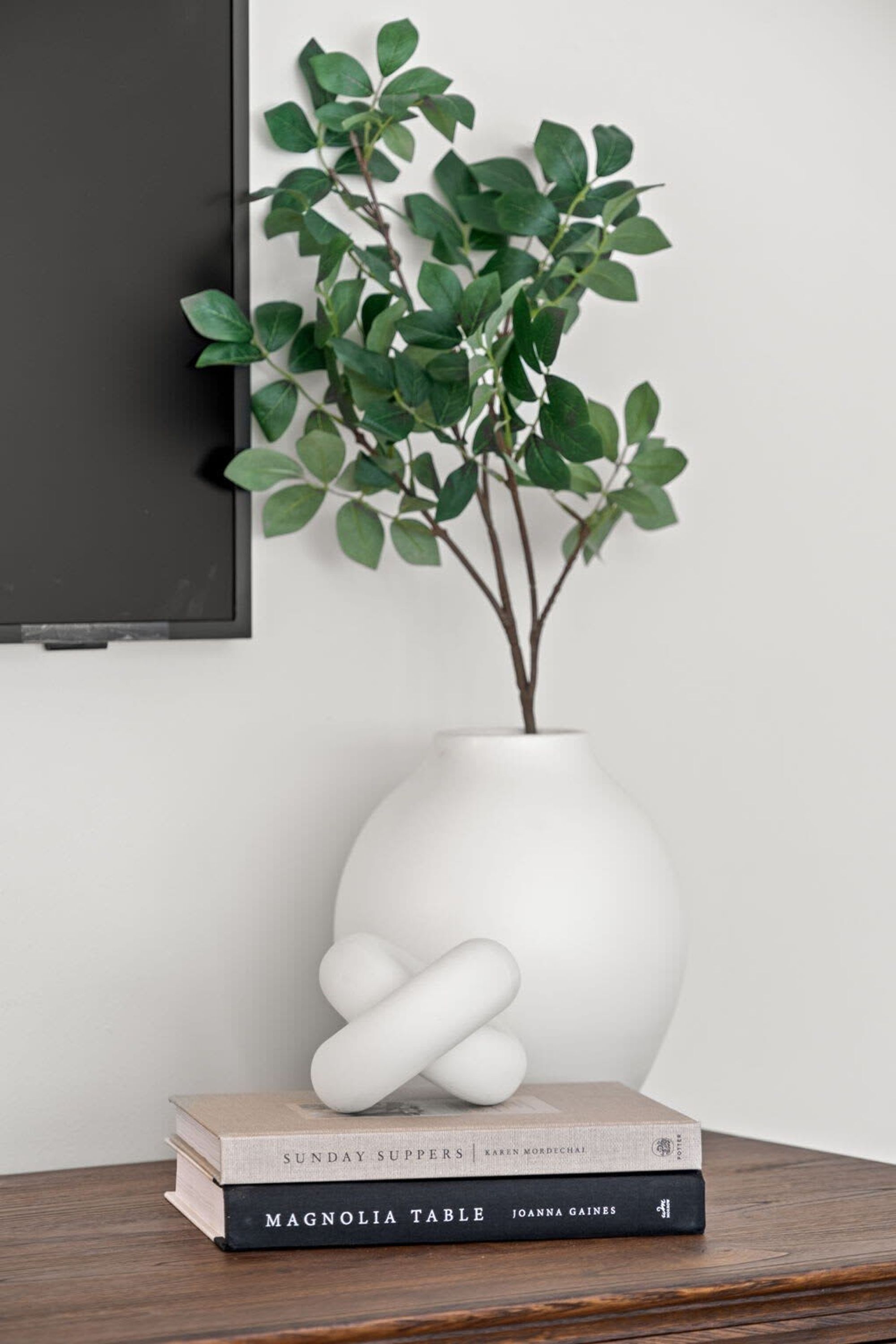 Eine weiße Vase mit einer Pflanze auf einem Holztisch, ein Buch mit dem Titel "Magnolia Table" von Joanna Gaines und ein gerahmtes Bild an der Wand.