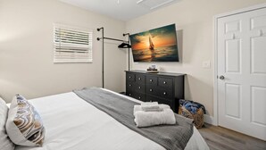 A bedroom with a bed, a dresser, a painting of a sunset over water, a window with blinds, and a door. - Soho Suite by Brightwild Steps to Soho (Tampa)