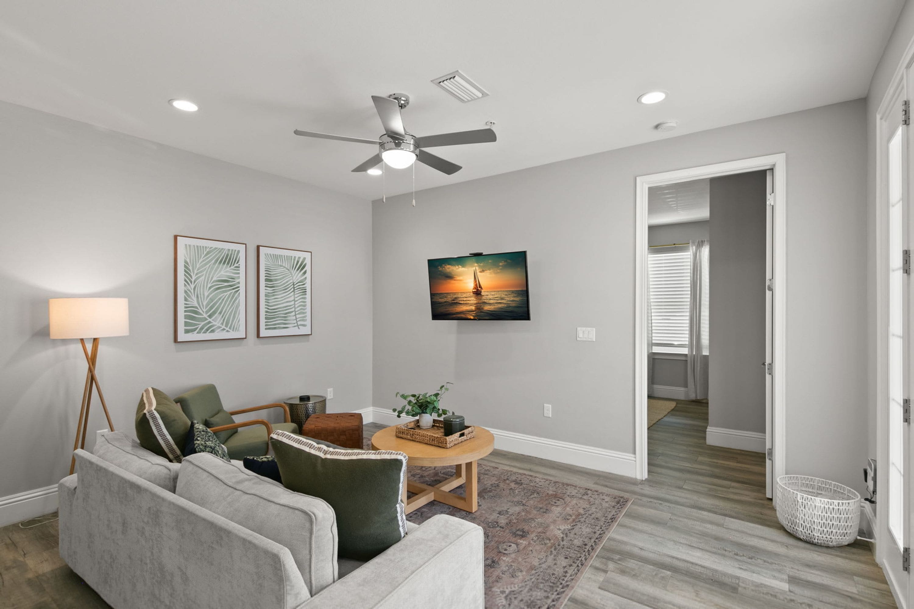 A modern living room with a grey sofa, a coffee table, and a ceiling fan. There is a painting on the wall and a door leading to another room.