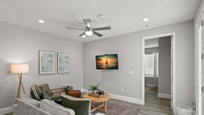 Un salon moderne avec un canapé gris, une table basse et un ventilateur de plafond. Un tableau est accroché au mur et une porte donne accès à une autre pièce.