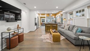 Ein modernes Wohnzimmer mit einer Couch, einem Hocker und einem Glastisch. Im Hintergrund ist eine Küchenzeile mit einer Theke und Barhocker zu sehen.