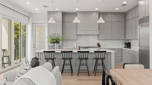 A modern kitchen with grey cabinets, a central island with bar stools, pendant lighting, and a large window providing natural light. - Jade House at The Gemma (Clearwater Beach)