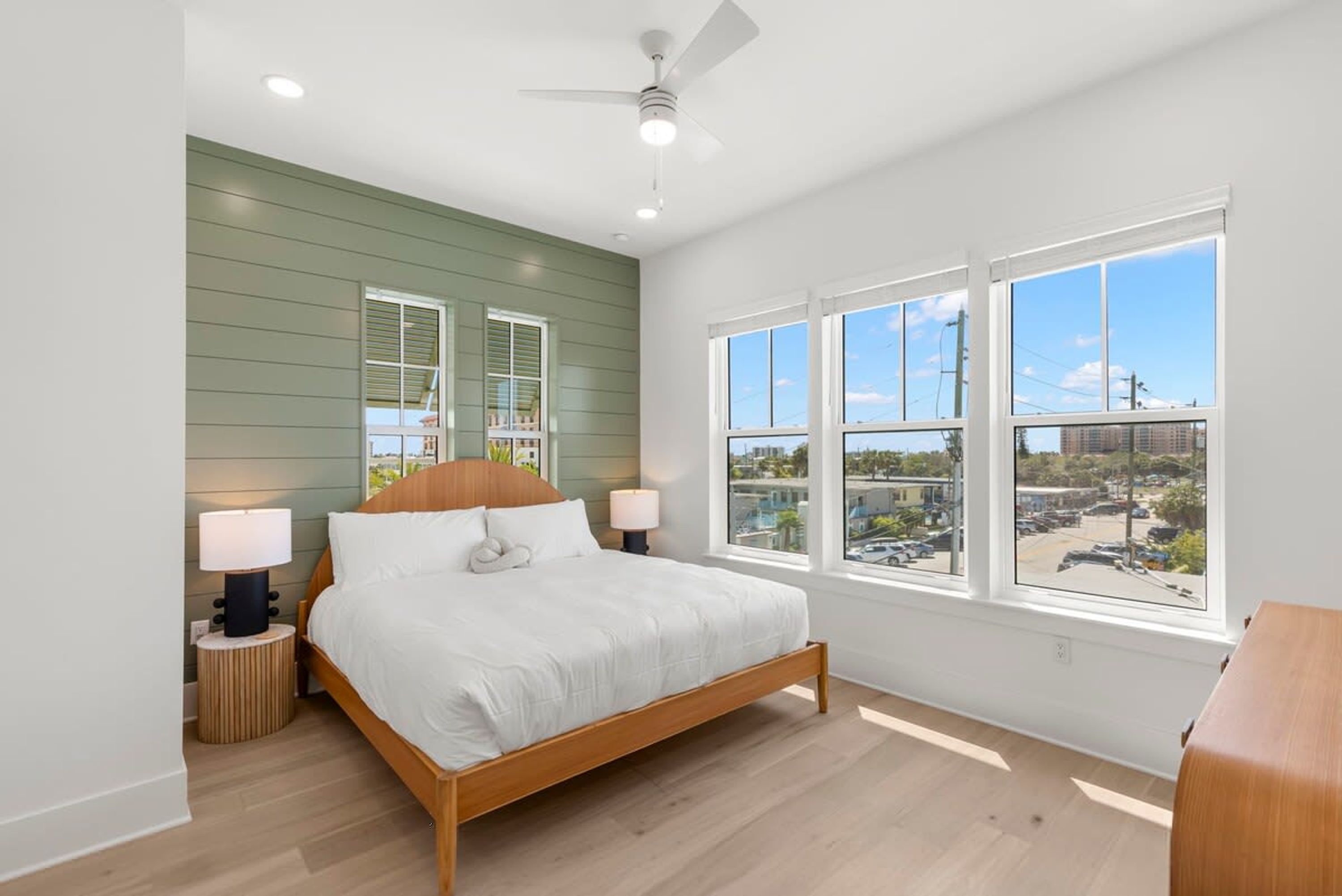 A bedroom with a bed, two bedside lamps, a ceiling fan, and large windows with a city view.