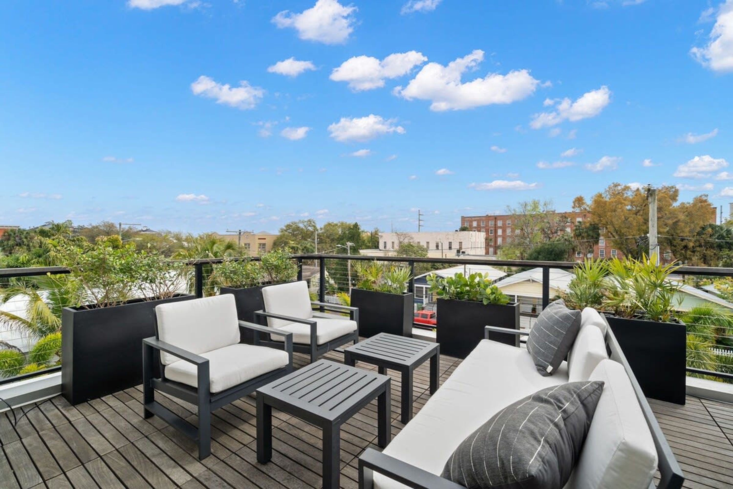 A rooftop terrace with outdoor seating, including a sofa and chairs, surrounded by greenery and overlooking a cityscape.