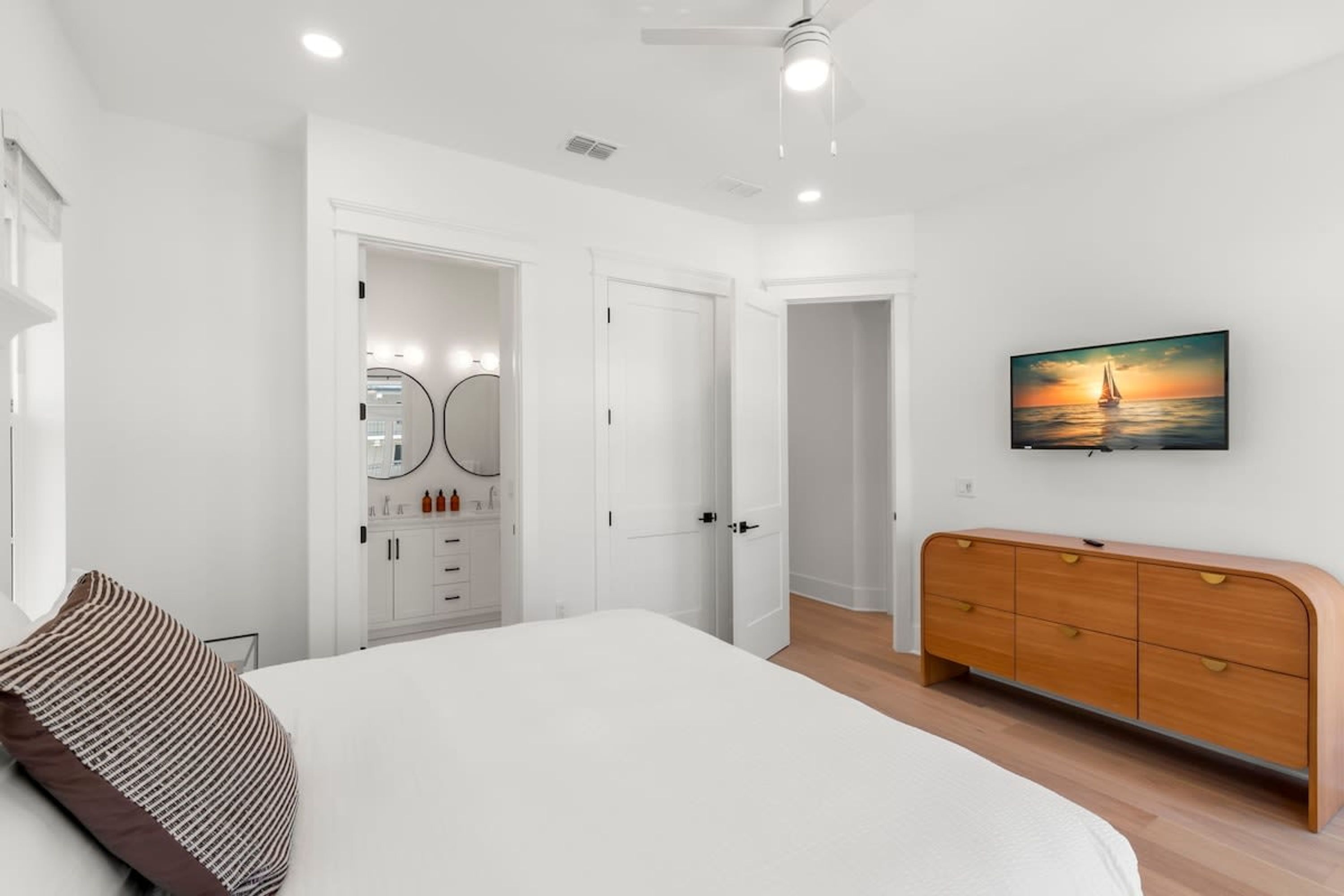 Quarto com cama, cômoda, televisão na parede e porta que dá para outro ambiente.