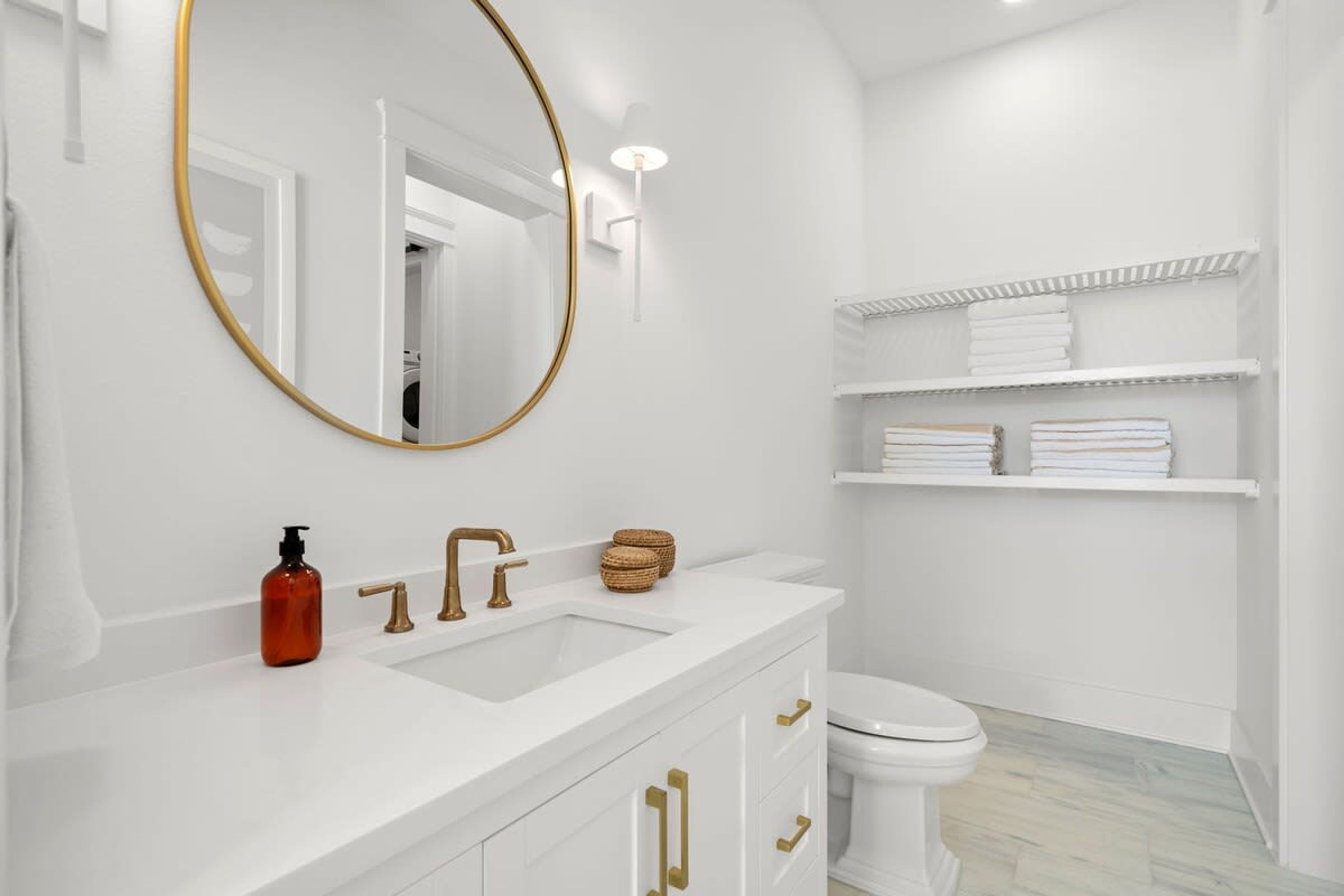 A modern bathroom with a white vanity, gold faucets, a round mirror, and white shelves with towels.