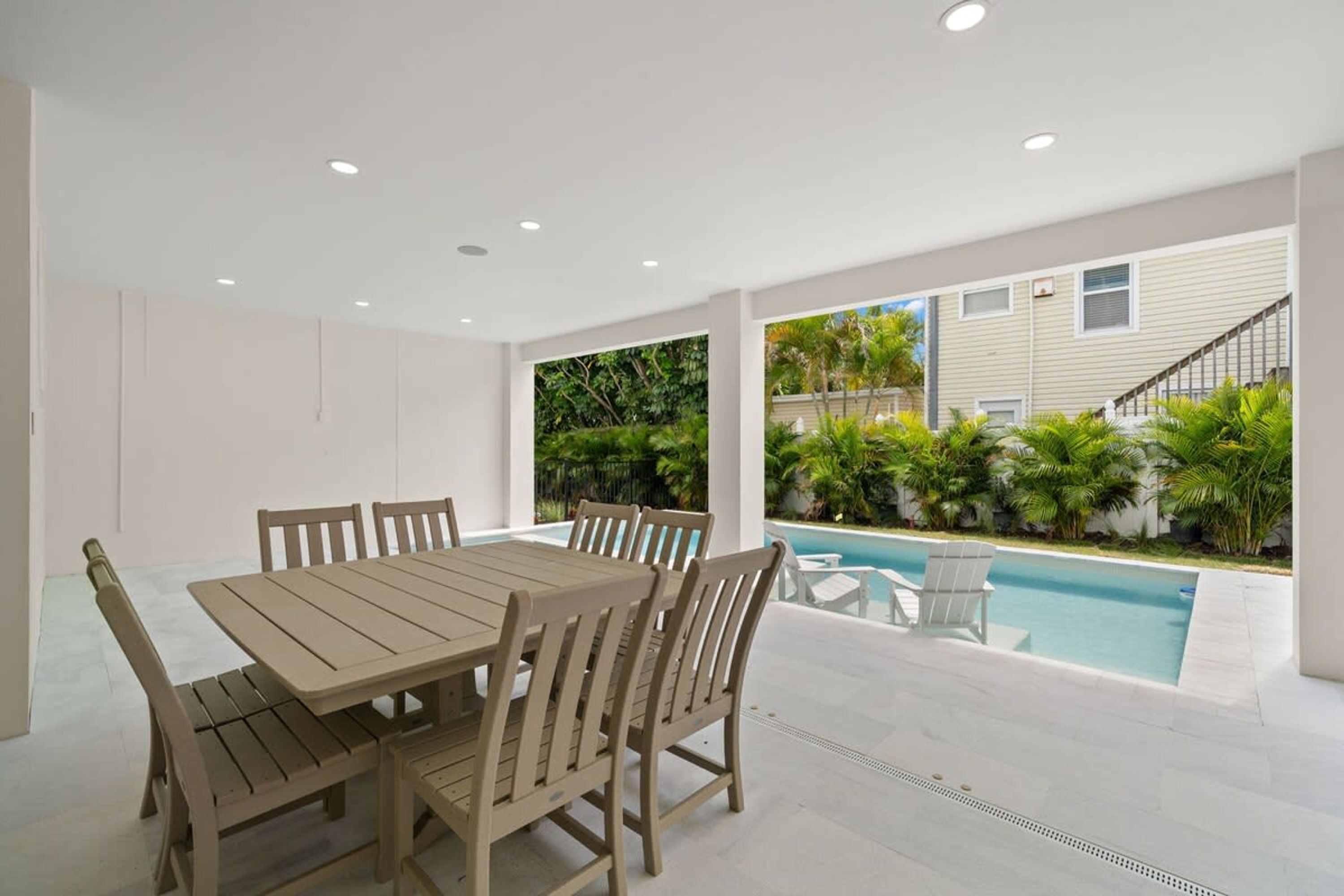 A spacious patio with a wooden table and chairs, a swimming pool, and a view of a residential area with palm trees.