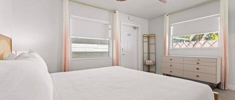 Un dormitorio con cama, ventilador de techo, ventana con cortinas, cómoda y puerta.