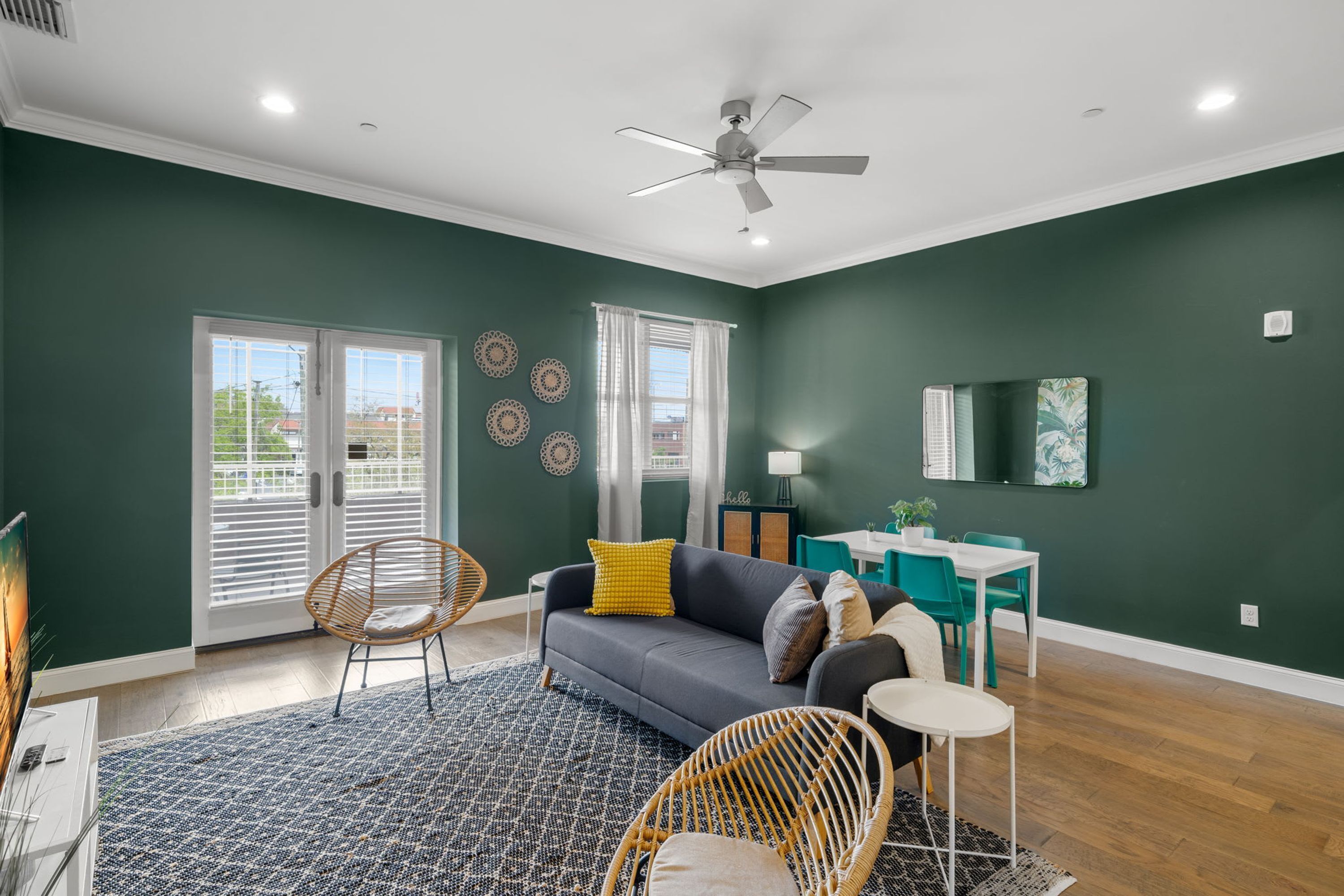 A living room with a dark green wall, a grey sofa, a patterned rug, wicker chairs, and a ceiling fan.