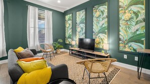 A living room with a sofa, chairs, a TV, and large tropical-themed paintings on the walls.
