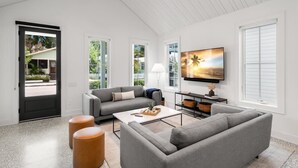 Un salon moderne avec un canapé gris, une table basse et un téléviseur fixé au mur. Une porte donne sur l’extérieur et la pièce est dotée de grandes fenêtres.