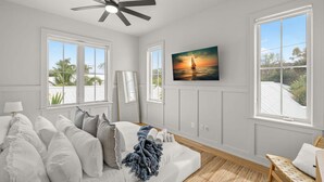 Une chambre à coucher avec un lit, un ventilateur de plafond, un tableau représentant un coucher de soleil et de grandes fenêtres.