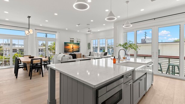 A modern kitchen with a large island, dining area, and large windows offering views of the city. - The Gemma Collection - 4 Stunning CWB Pool Homes (Clearwater Beach)