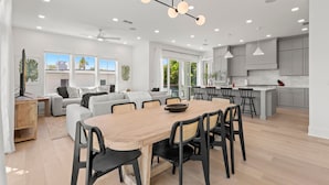 A modern kitchen with white cabinets, a central island, and a dining area with a wooden table and chairs. - The Gemma Collection - 4 Stunning CWB Pool Homes (Clearwater Beach)
