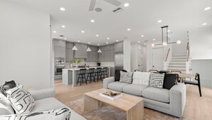 A modern living room with a grey sofa, a wooden coffee table, and a kitchen in the background. - The Gemma Collection - 4 Stunning CWB Pool Homes (Clearwater Beach)