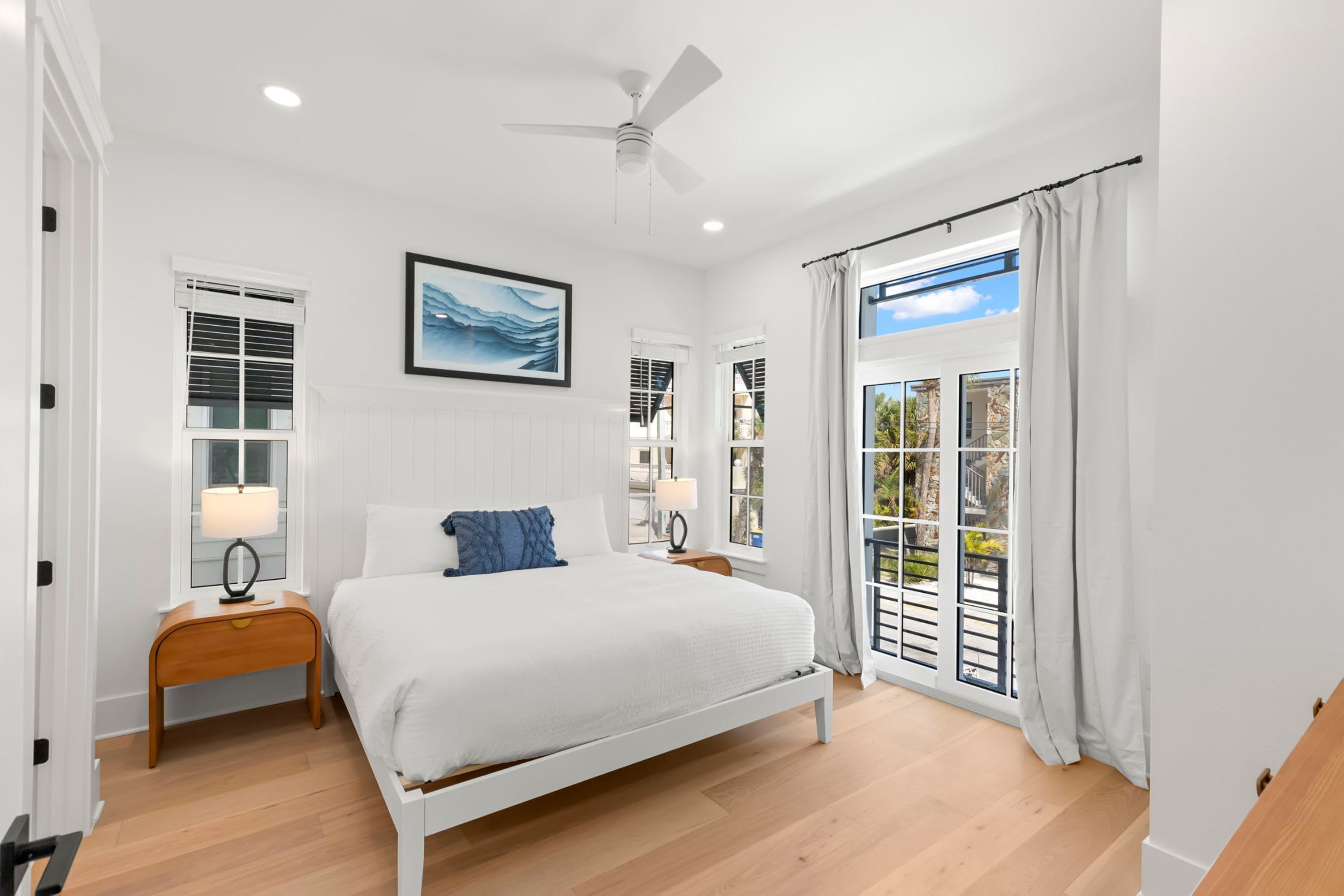 Une chambre avec un lit blanc, des tables de chevet en bois, un ventilateur de plafond et une vue sur l’extérieur, accessible par une porte-fenêtre coulissante.